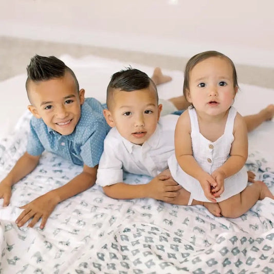 Three children sitting on an Eclectic Aden + Anais Now + Zen cotton muslin blanket with a neutral koala print.