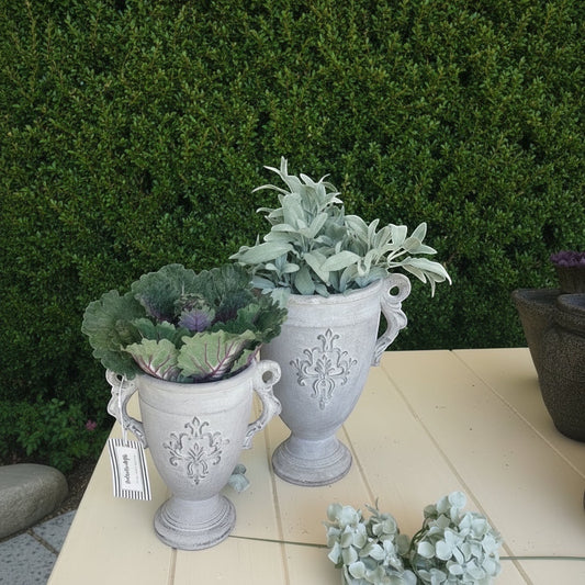 Eclectic antique cement trophy vase planters with ornate handles and weathered finish on a table with greenery