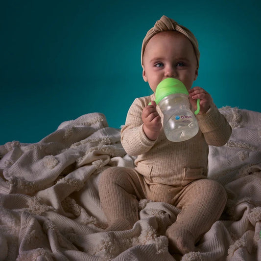 Eclectic b.box glow in the dark sippy cup with weighted straw held by a baby sitting on a textured cream blanket