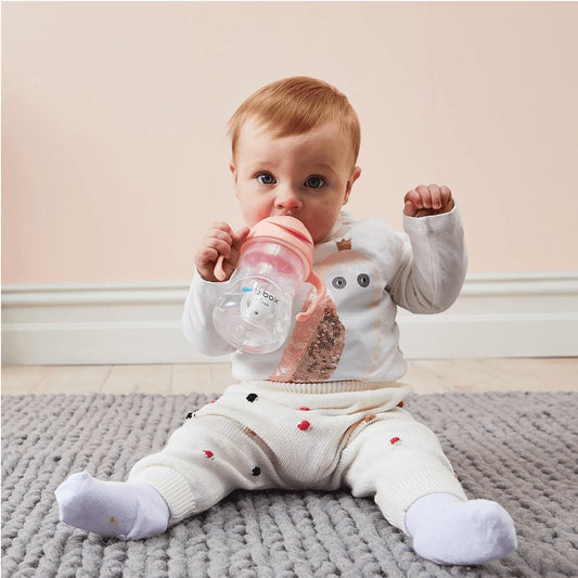 Eclectic b.box Tutti Frutti weighted straw sippy cup held by a baby sitting on a grey rug