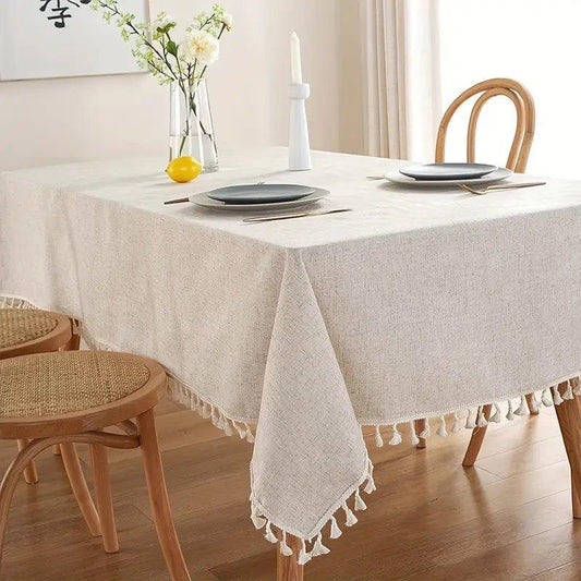 Eclectic light beige linen blend rectangular tablecloth with decorative tassel trim on a wooden dining table.