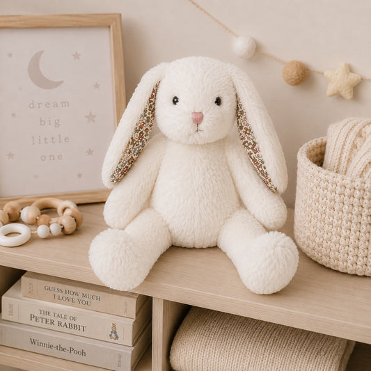 Eclectic Bun-Bun white plush bunny toy with floral ear lining sitting on a wooden nursery shelf with books.
