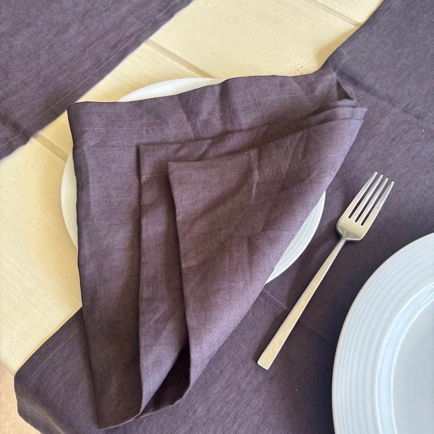 Eclectic charcoal linen table runner and matching napkin set on a light wooden table with white plates and a fork.