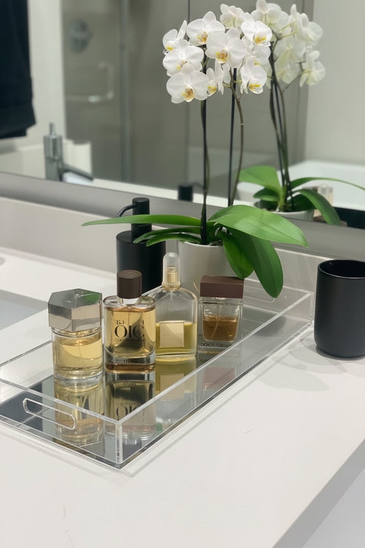 Eclectic clear acrylic mirrored display tray holding luxury perfume bottles on a bathroom vanity with an orchid plant.