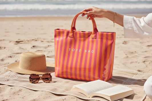 Eclectic coral and pink striped PVC tote bag held on a sandy beach with a straw hat, sunglasses, and an open book.