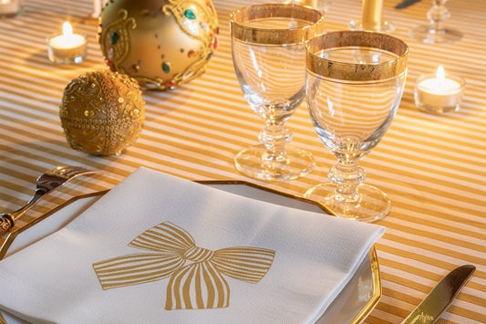 Eclectic Françoise Paviot Noeud Or cocktail napkin with gold bow design on a striped table setting with glassware