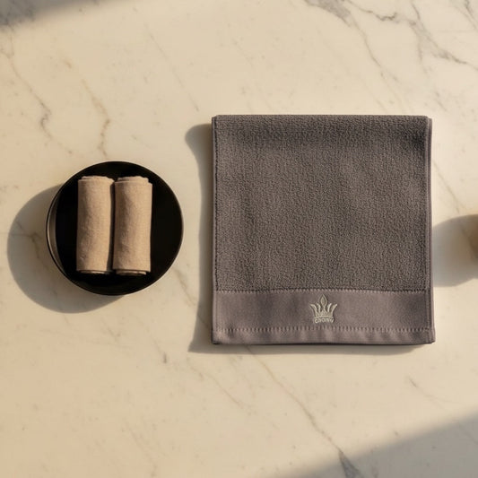 Eclectic grey cotton guest towel with embroidered crown detail on a marble surface next to rolled towels.