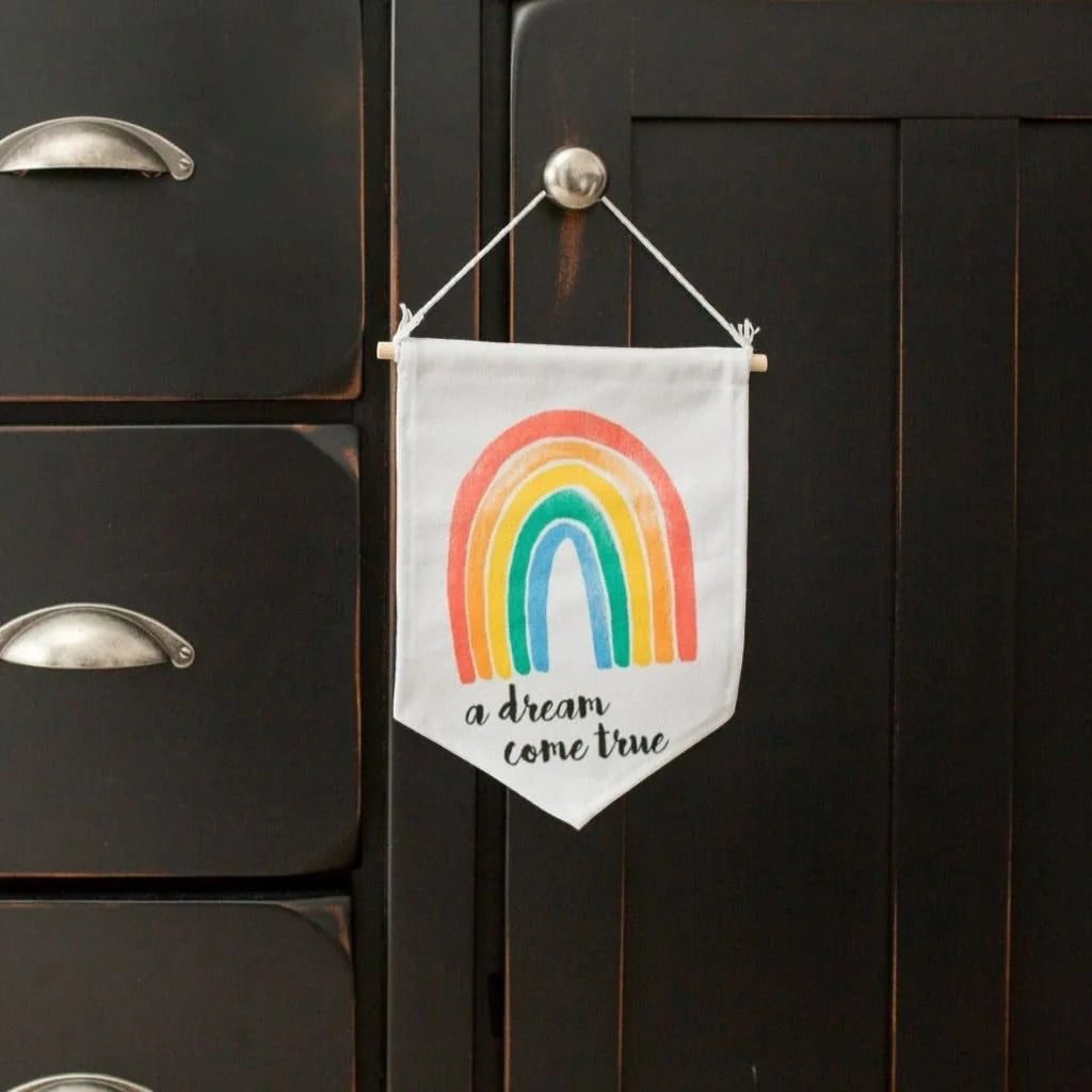 Eclectic Lulujo rainbow sky nursery wall banner with wooden hanger hanging on a dark cabinet
