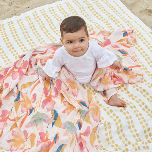 Eclectic baby sitting on a soft Marine Garden bamboo muslin swaddle blanket with floral and dotted patterns on sand.