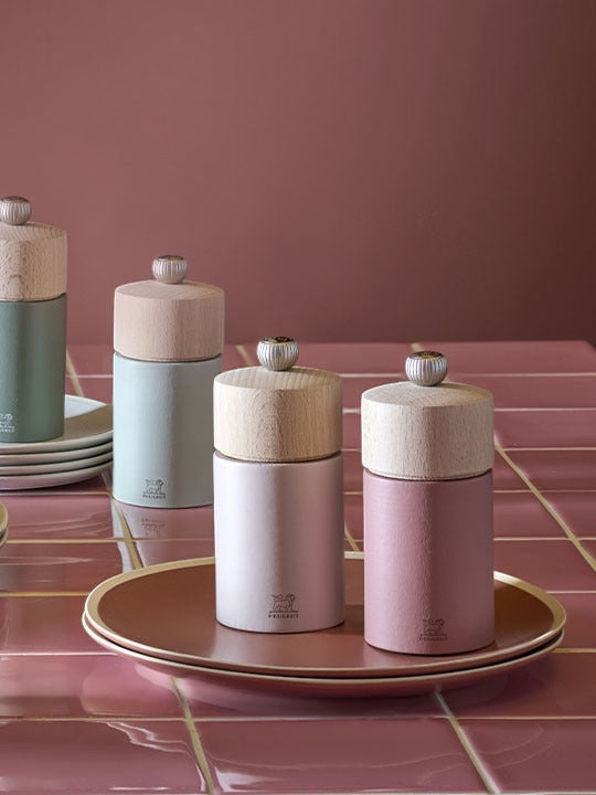 Eclectic Peugeot Boreal salt and pepper mills in sage green and pink on a tiled table with matching plates.