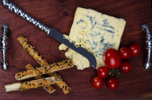 Eclectic Ribbon pewter cheese knife with a vine-inspired handle, served on a wooden board with blue cheese and tomatoes.