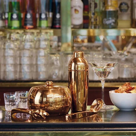 Eclectic Sambonet gold PVD stainless steel ice spoon with perforated bowl on a bar counter with cocktail accessories