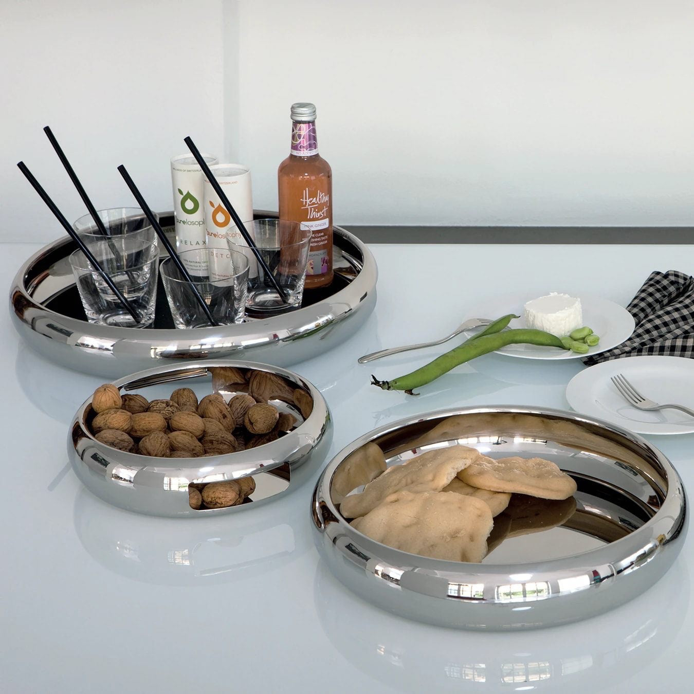 Eclectic Sambonet Sphera mirror-polished stainless steel serving bowls arranged on a white table with snacks and drinks.