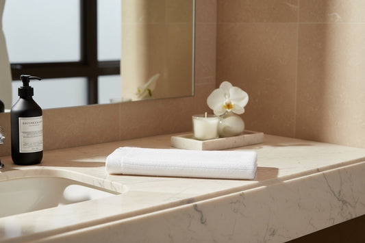 Eclectic white velvet cotton guest towel folded on a marble bathroom vanity with a candle and soap dispenser.