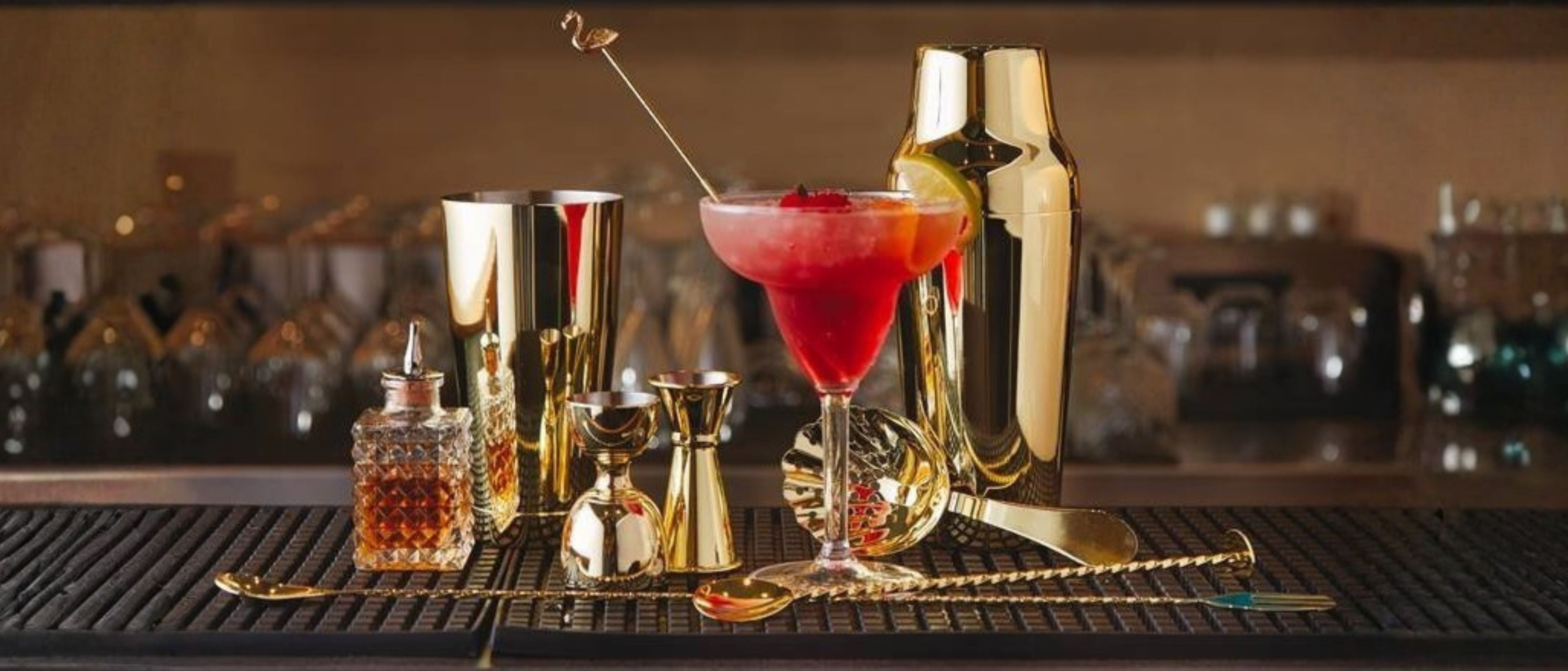 Gold bar accessories with cocktail shaker, jigger, strainer, stirrer, and cocktail glass on a bar counter.