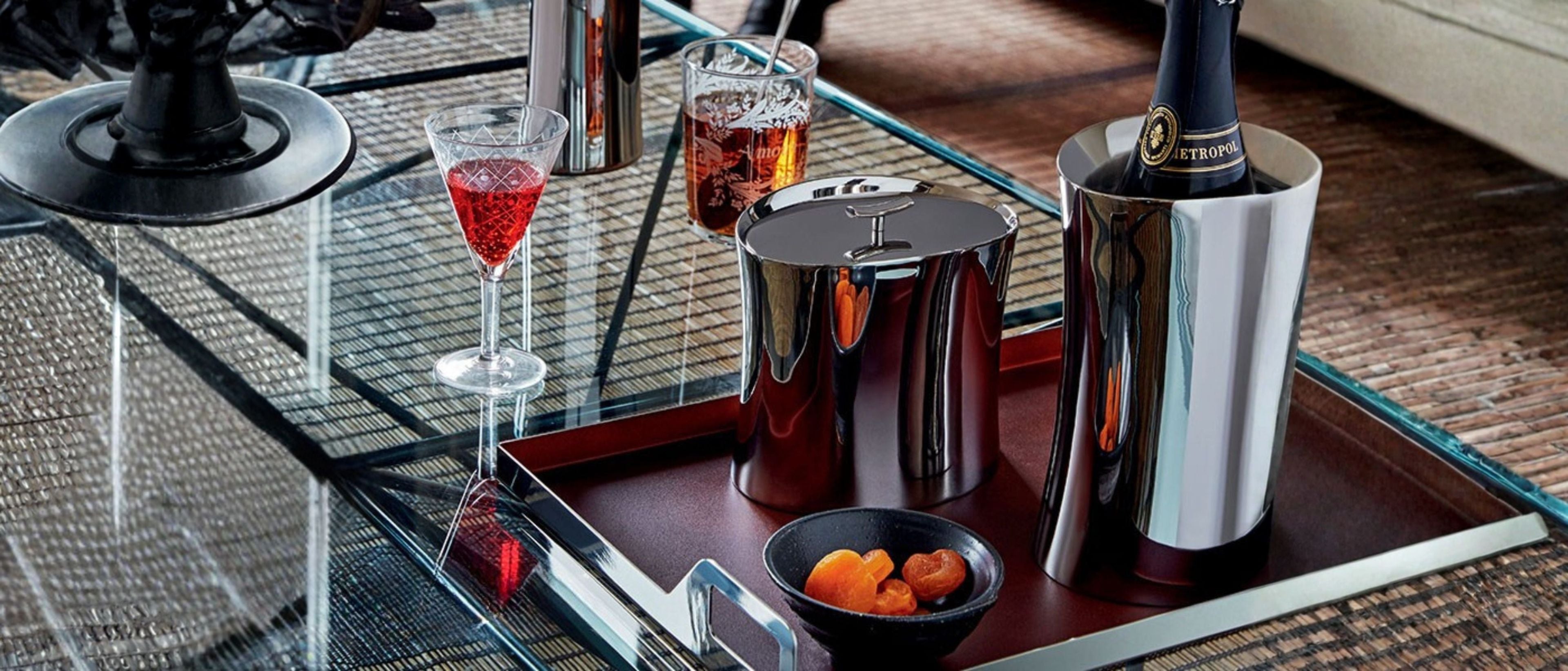 Barware collection with glasses, ice bucket, bottle cooler, tray, and cocktail accessories on a glass table.