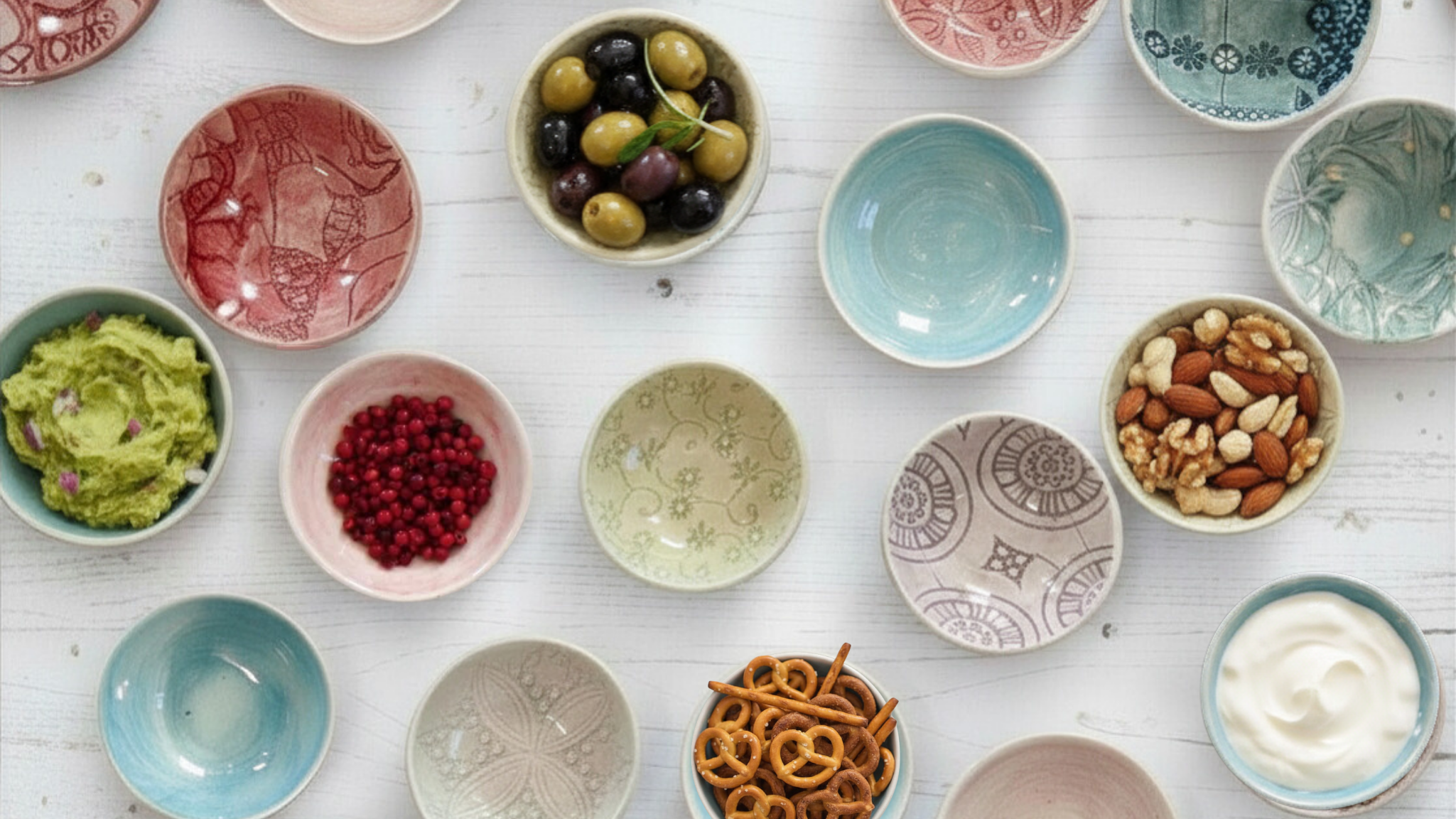 Colourful condiment and small bowls filled with olives, nuts, dips, and snacks on a white table.
