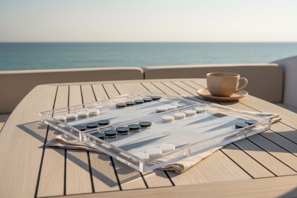 Eclectic modern clear acrylic backgammon set with black and white playing pieces on a wooden table by the sea.