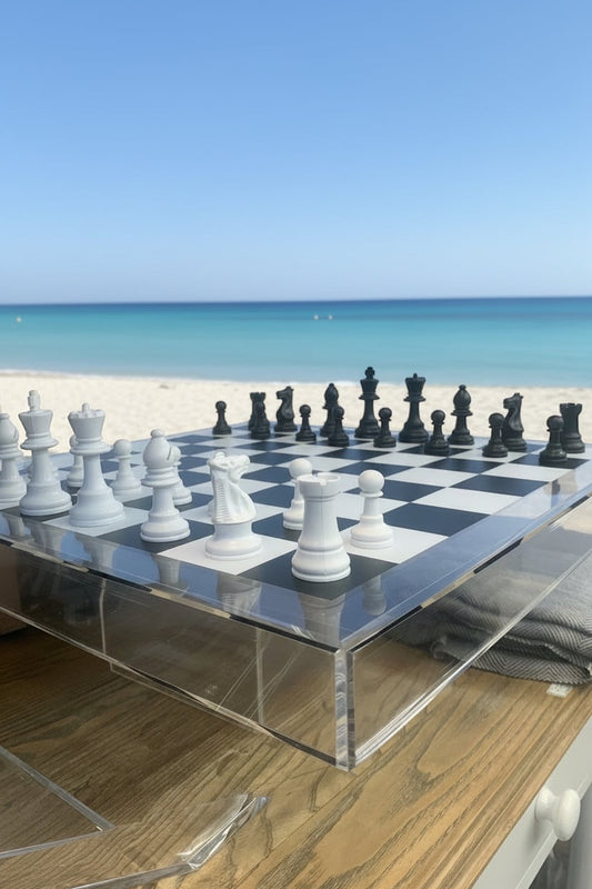 Eclectic 56cm acrylic chess board with black and white pieces set on a wooden table overlooking a sunny beach.