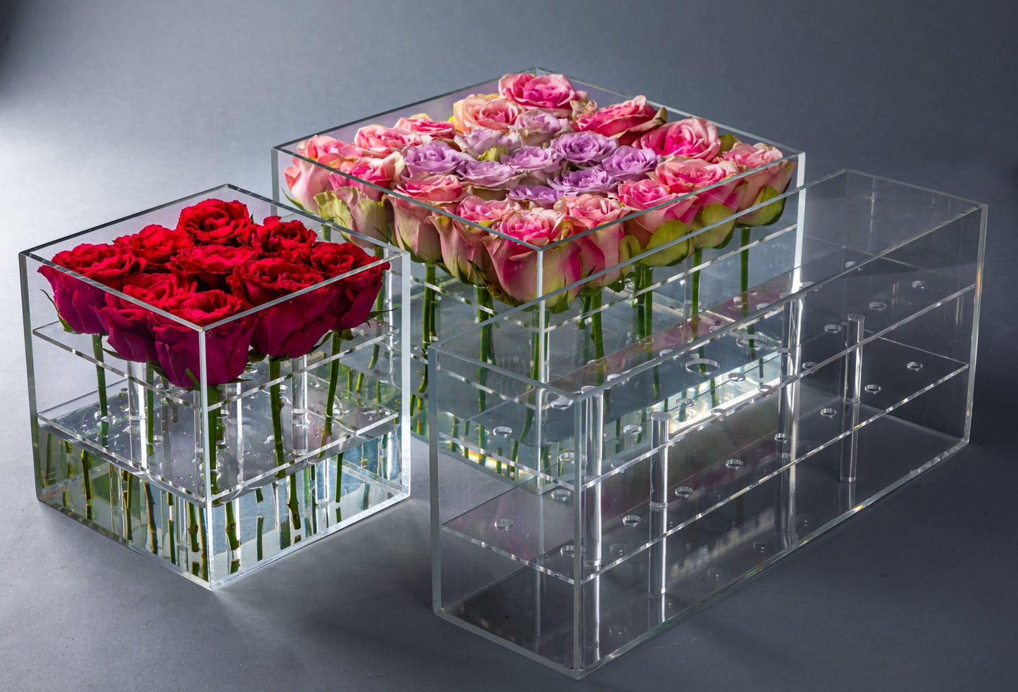 Eclectic rose box set featuring clear acrylic containers filled with vibrant red and pink roses on a grey background.