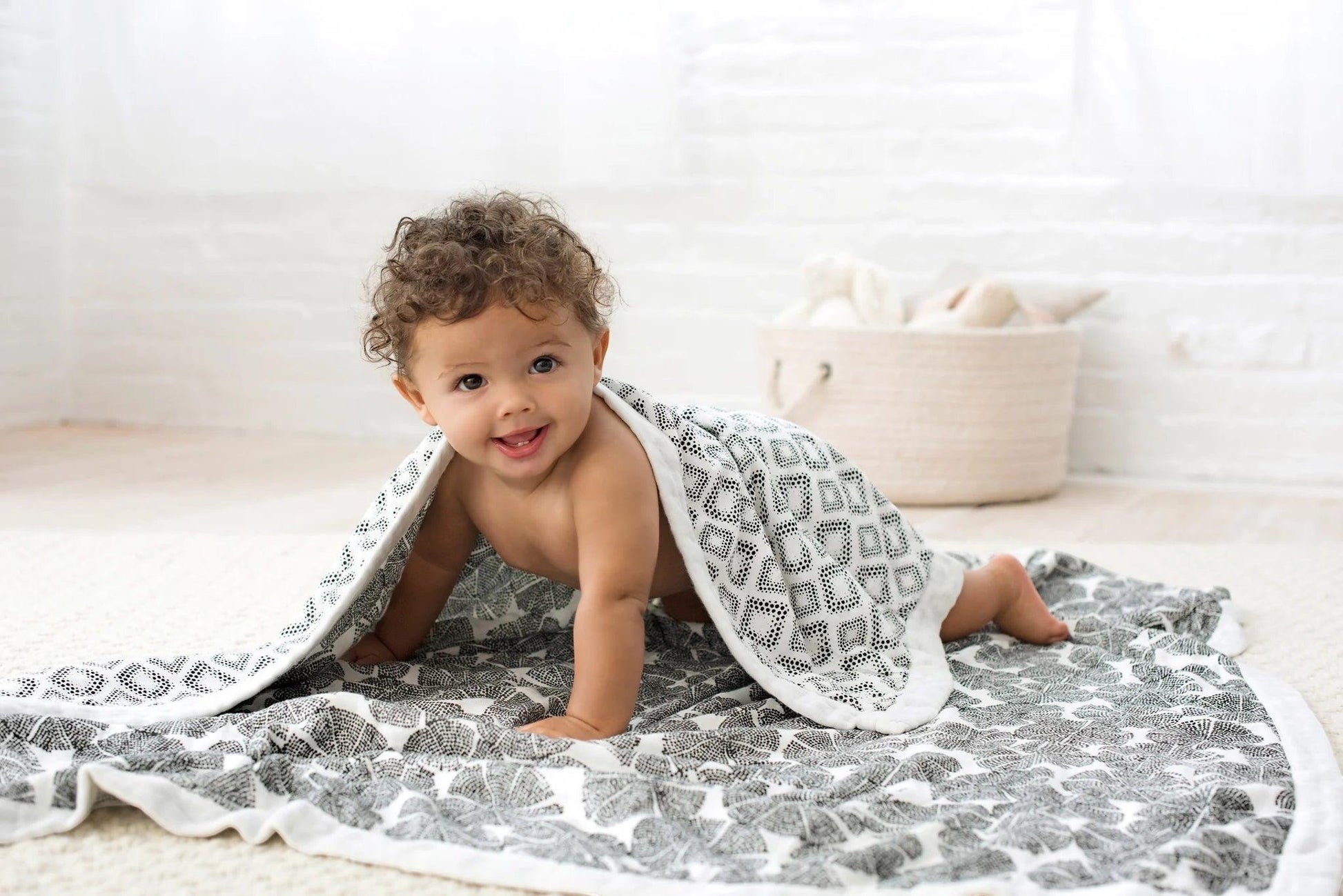 Eclectic Aden + Anais In Motion Wedges bamboo muslin baby blanket draped over a smiling baby on a nursery floor