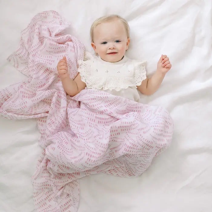 Eclectic Aden + Anais Deco muslin swaddle blanket in pink draped over a baby lying on a white sheet