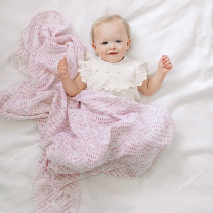 Eclectic Aden + Anais Deco muslin swaddle blanket in pink draped over a baby lying on a white sheet