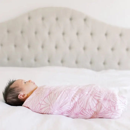 Eclectic Aden + Anais baby swaddled in a pink and white Deco print cotton muslin blanket on a bed