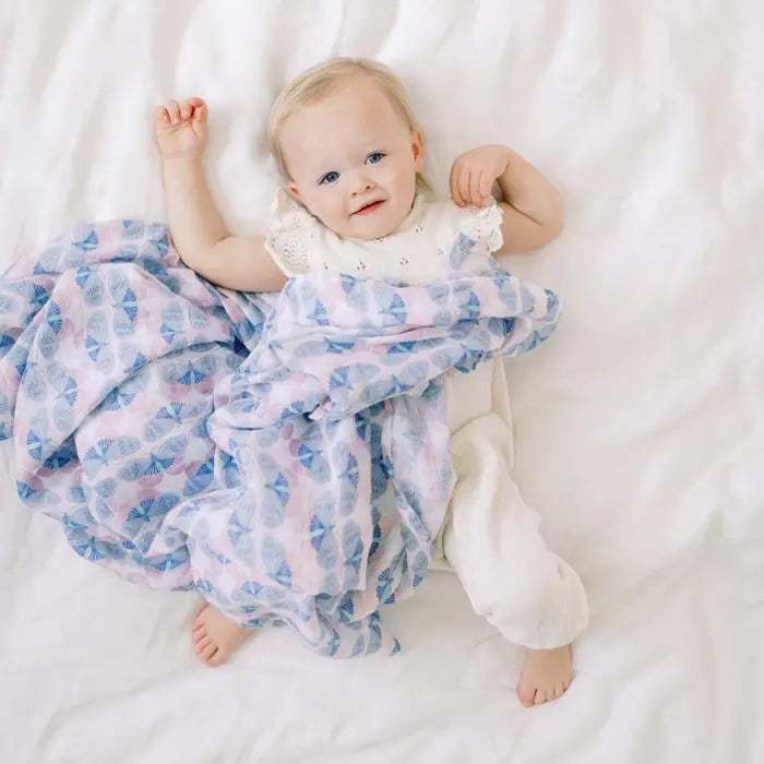 Baby lying on a white sheet covered by an Aden + Anais cotton muslin swaddle with a blue and pink Deco print