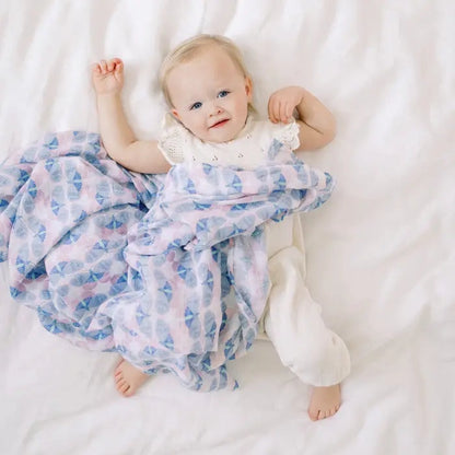 Baby lying on a white sheet covered by an Aden + Anais cotton muslin swaddle with a blue and pink Deco print