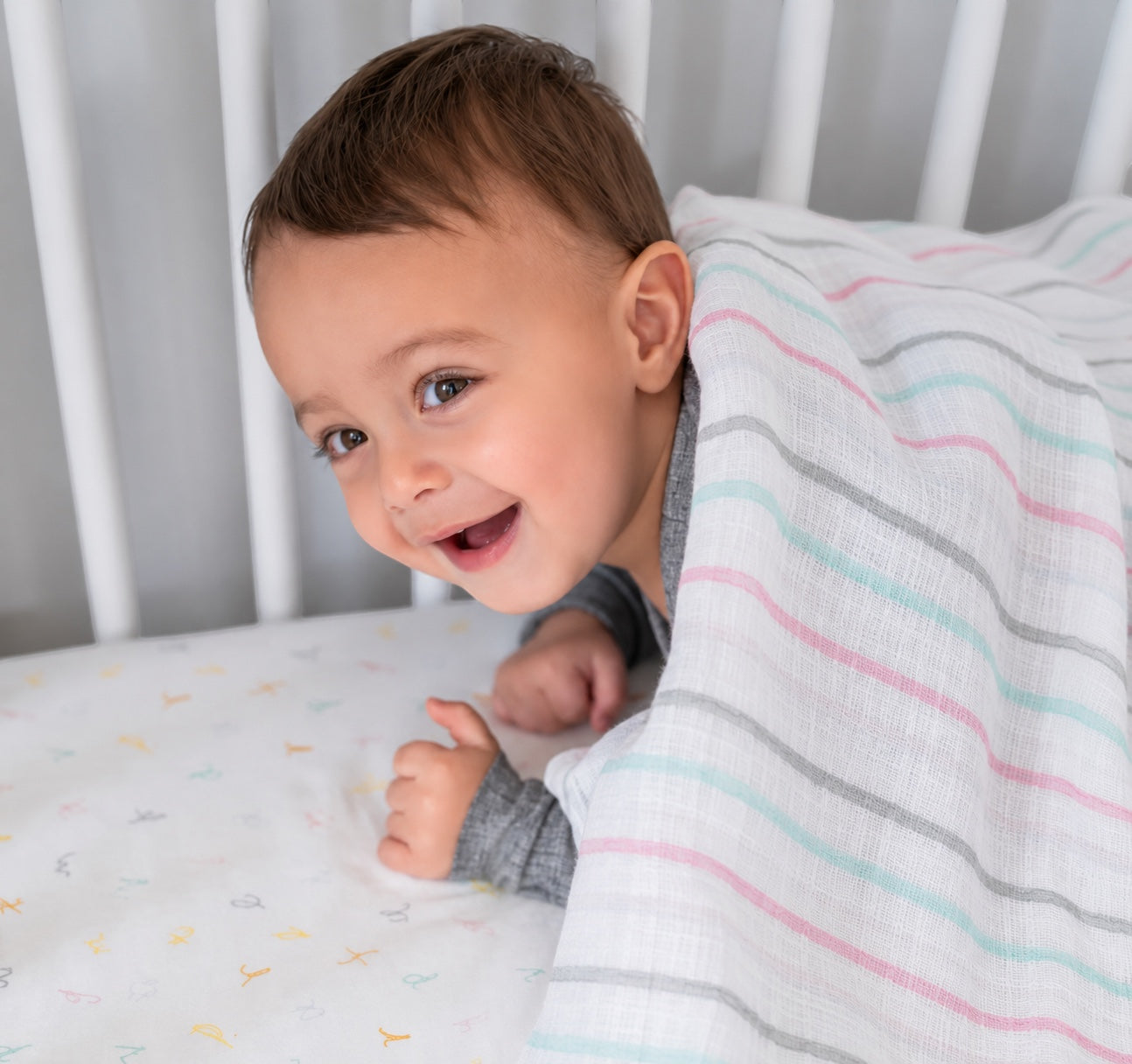 Eclectic Aden + Anais pastel stripe muslin swaddle blanket draped over a smiling baby in a white crib