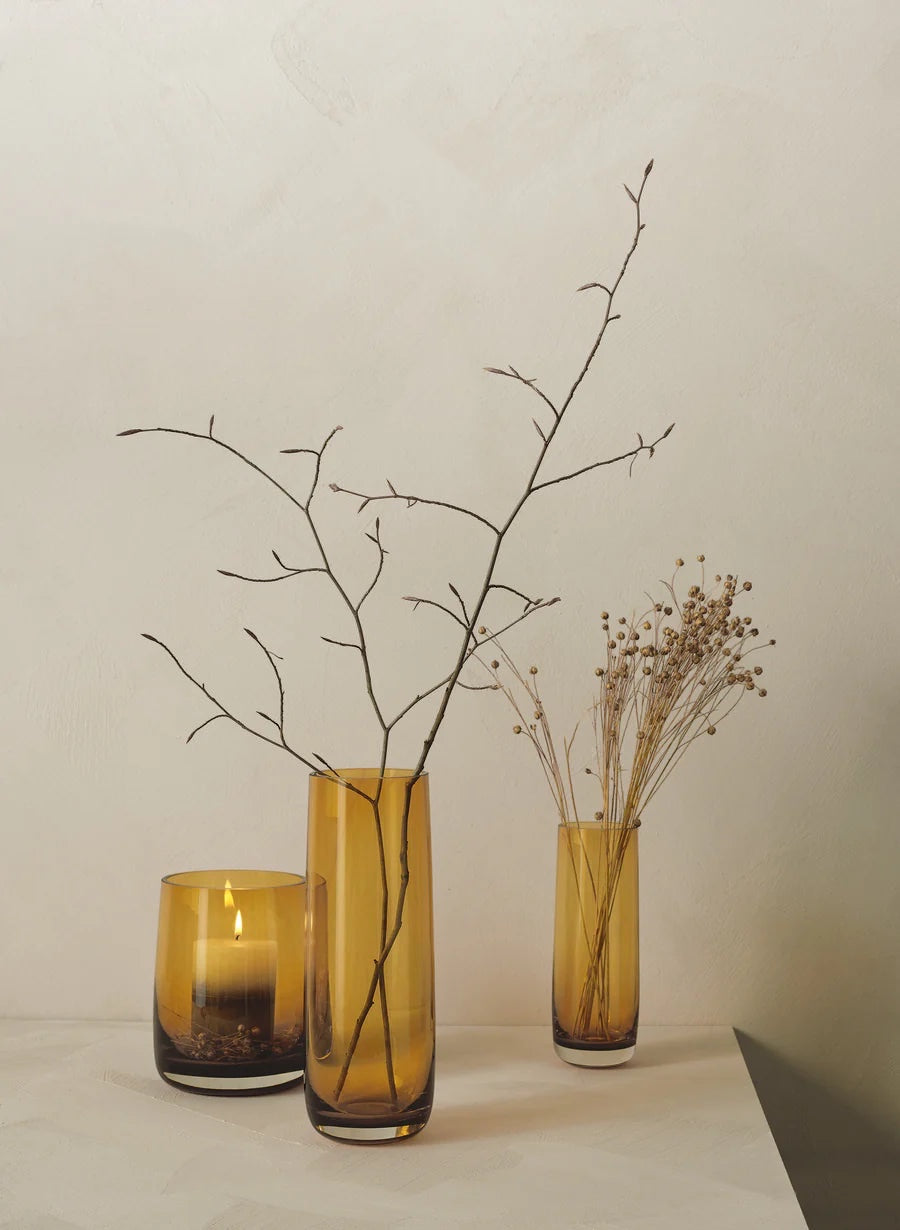 Eclectic Ajana amber mouth-blown glass vases and candle holder with dried branches on a neutral surface.