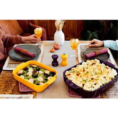 Eclectic Appolia ceramic oven dishes in saffron yellow and purple on a rustic dining table with food and drinks.