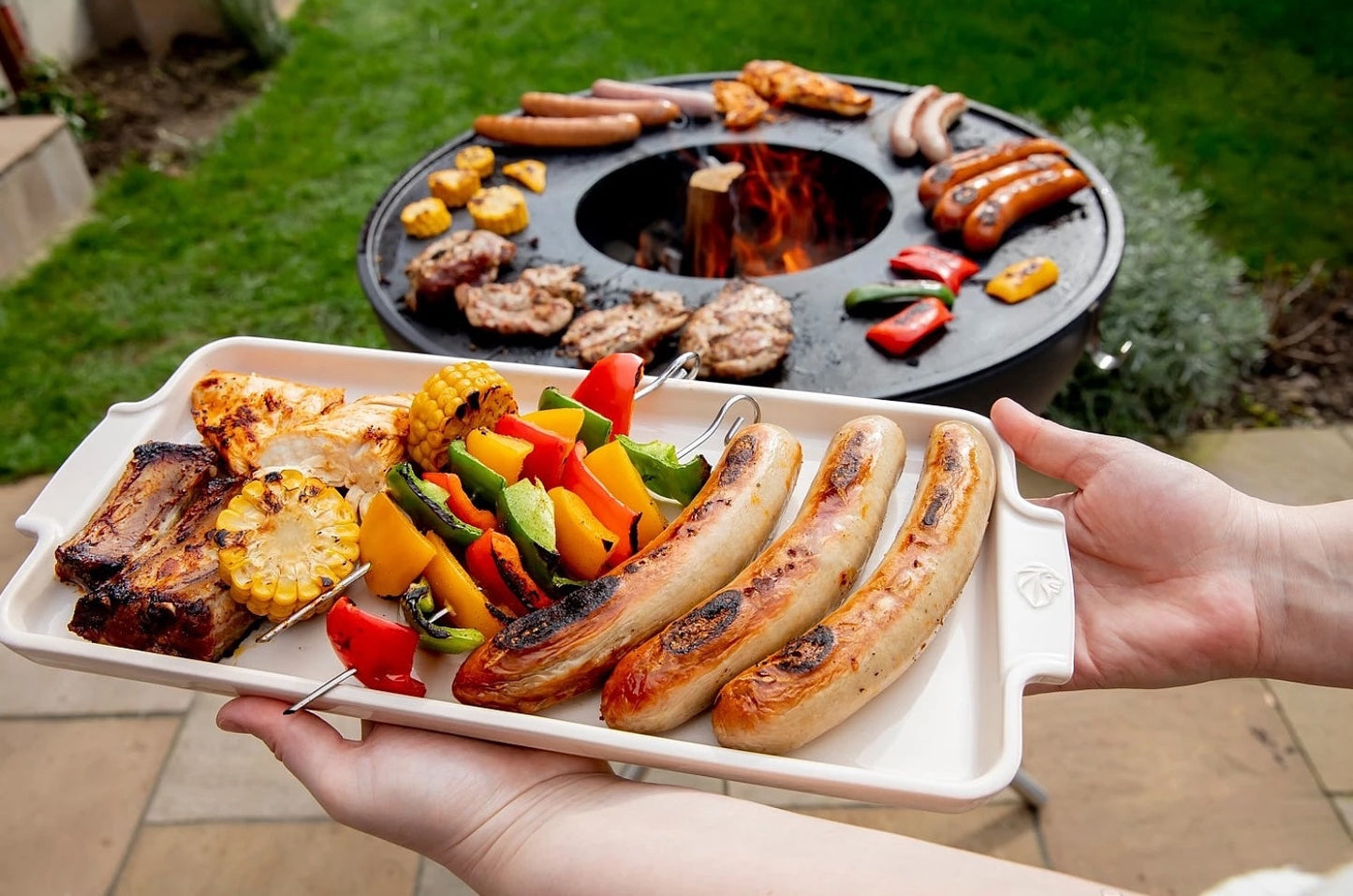 Eclectic Appolia ecru ceramic appetizer platter held by hands, filled with grilled sausages and vegetables outdoors.