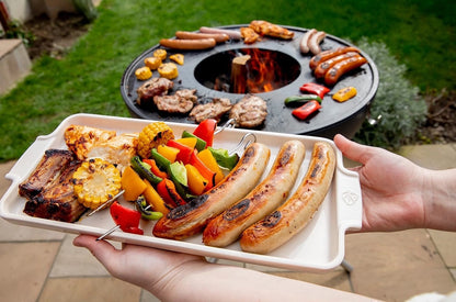Eclectic Appolia ecru ceramic appetizer platter held by hands, filled with grilled sausages and vegetables outdoors.