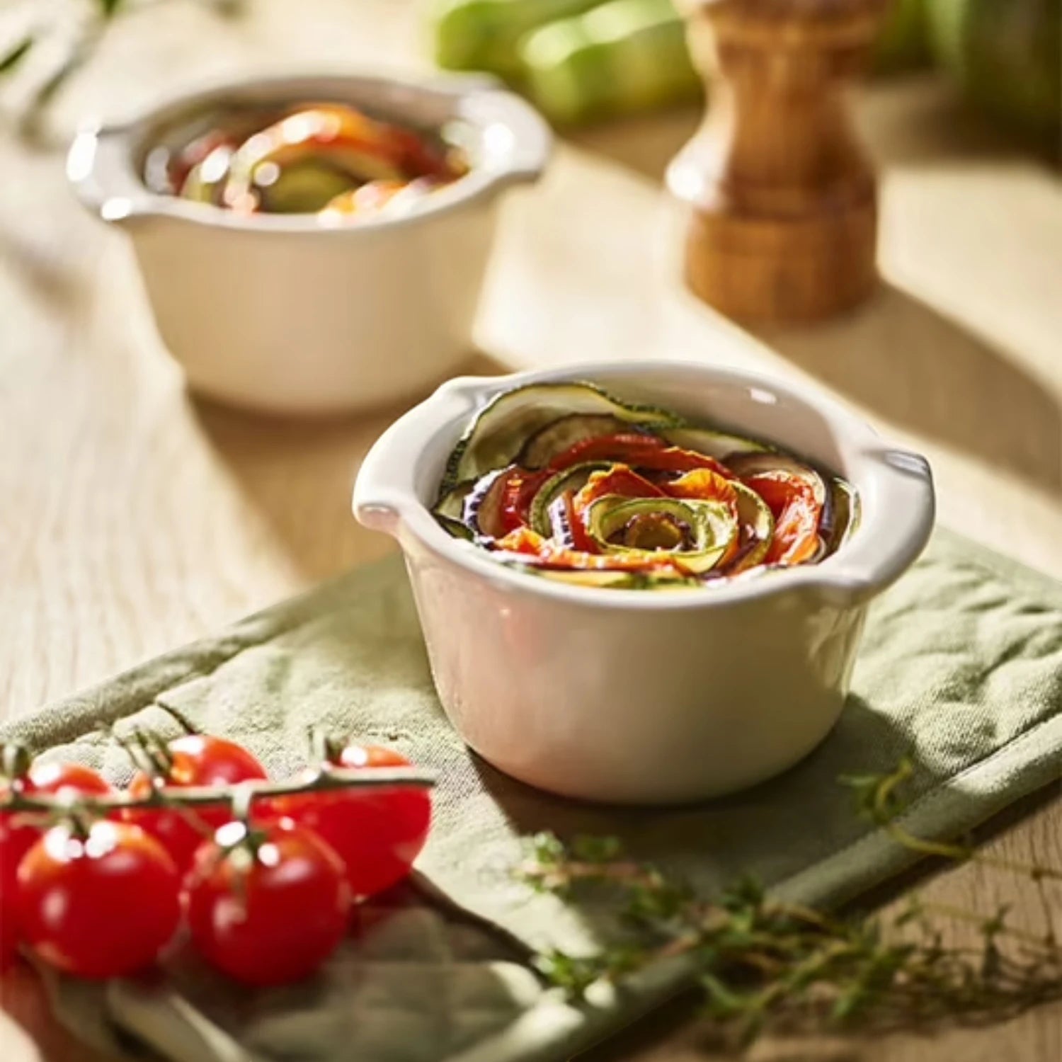 Eclectic Appolia ecru ceramic ramekins filled with baked vegetable tian on a wooden table with cherry tomatoes.