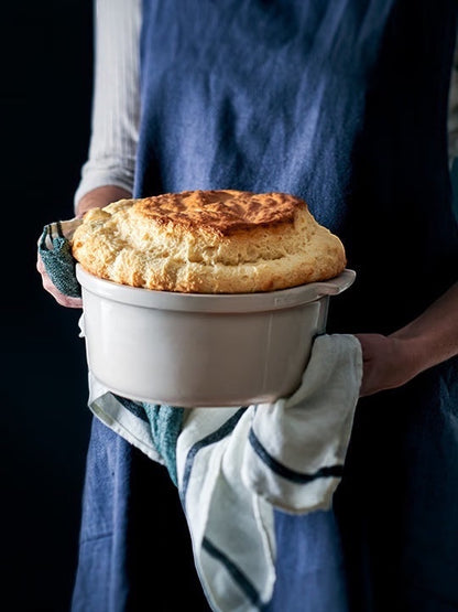 Eclectic Appolia 24 cm ecru ceramic souffle dish held by a person in a blue apron with a freshly baked souffle