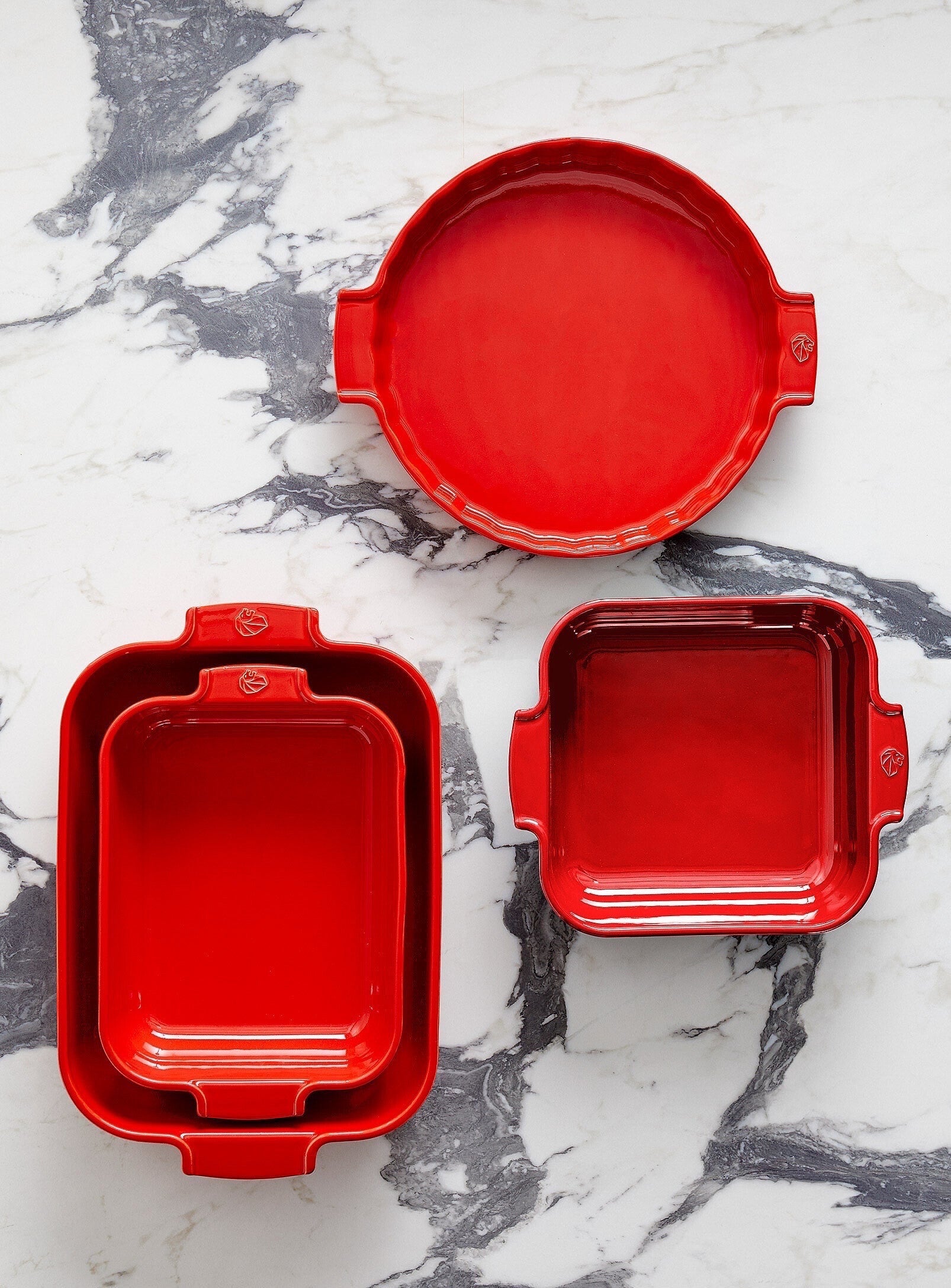 Eclectic Appolia red ceramic oven dishes in various shapes and sizes arranged on a white marble surface.