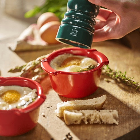 Eclectic Appolia rich red ceramic ramekins holding baked eggs with a green pepper mill on a wooden kitchen board