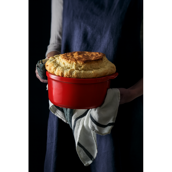 Eclectic Appolia 24 cm red ceramic soufflé dish held by a person in a blue apron with a freshly baked golden soufflé.