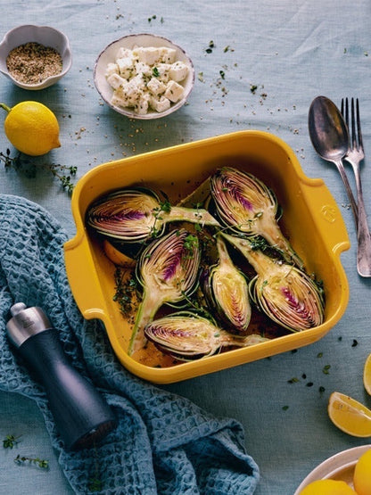 Eclectic Appolia saffron yellow ceramic square oven dish filled with roasted artichokes on a blue linen tablecloth.