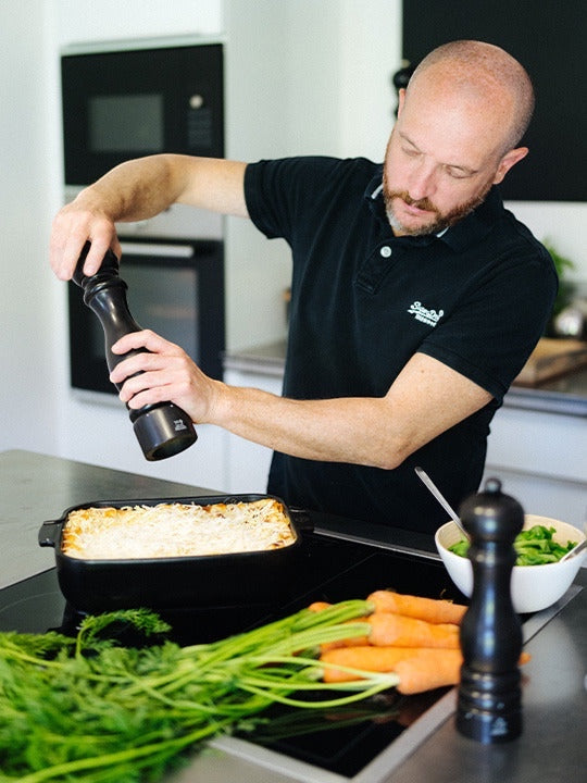 Eclectic Appolia satin black ceramic oven dish with a baked meal being seasoned by a man in a modern kitchen