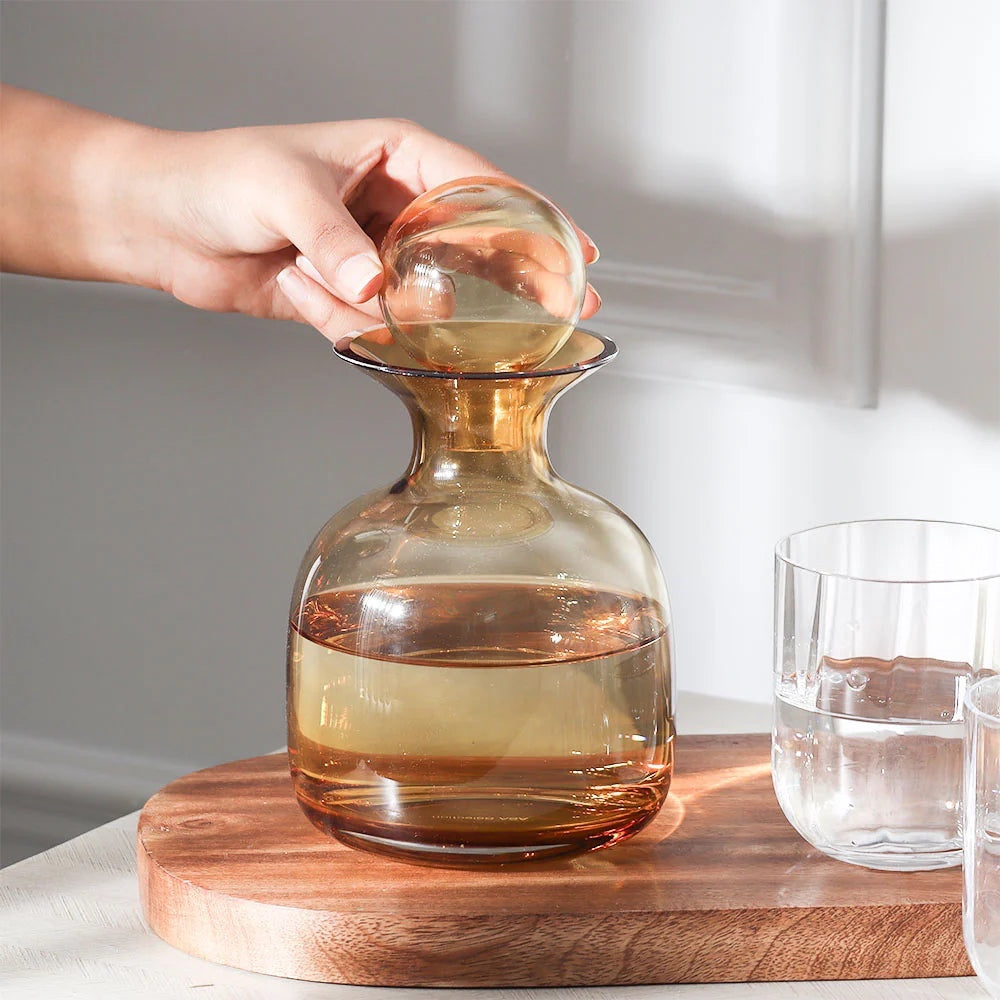 Eclectic ASA Selection Sarabi amber glass carafe with a spherical stopper being placed on a wooden serving board.