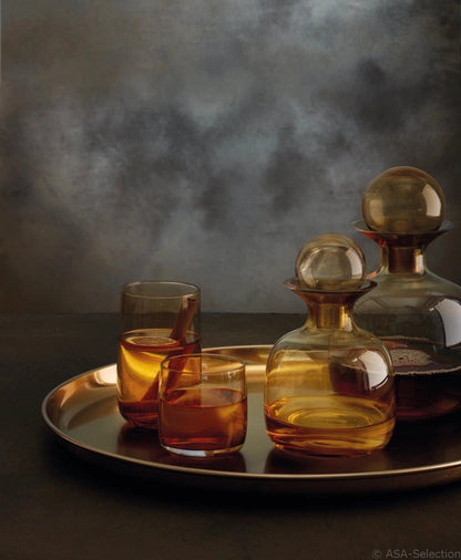 Eclectic ASA Selection Sarabi amber glass carafes and matching tumblers on a gold tray against a dark moody background.