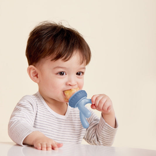 Eclectic B.Box blue silicone fresh food feeder held by a toddler in a striped shirt for safe self-feeding