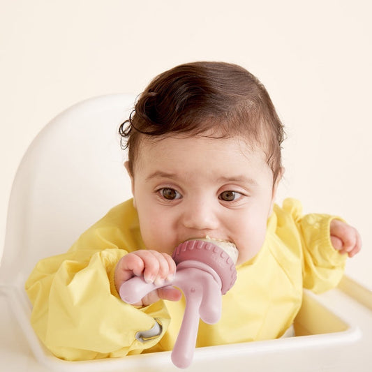 Eclectic B.Box blush silicone fresh food feeder held by a baby in a yellow bib sitting in a high chair