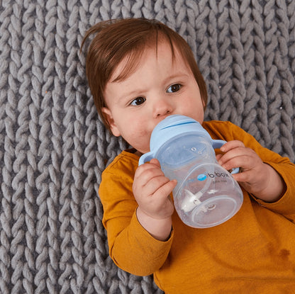 Eclectic b.box bubblegum sippy cup with weighted straw held by a baby lying on a grey chunky knit blanket