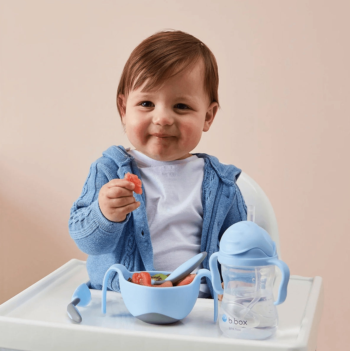 Eclectic toddler sitting in a high chair with a b.box bubblegum sippy cup and matching blue bowl of fruit