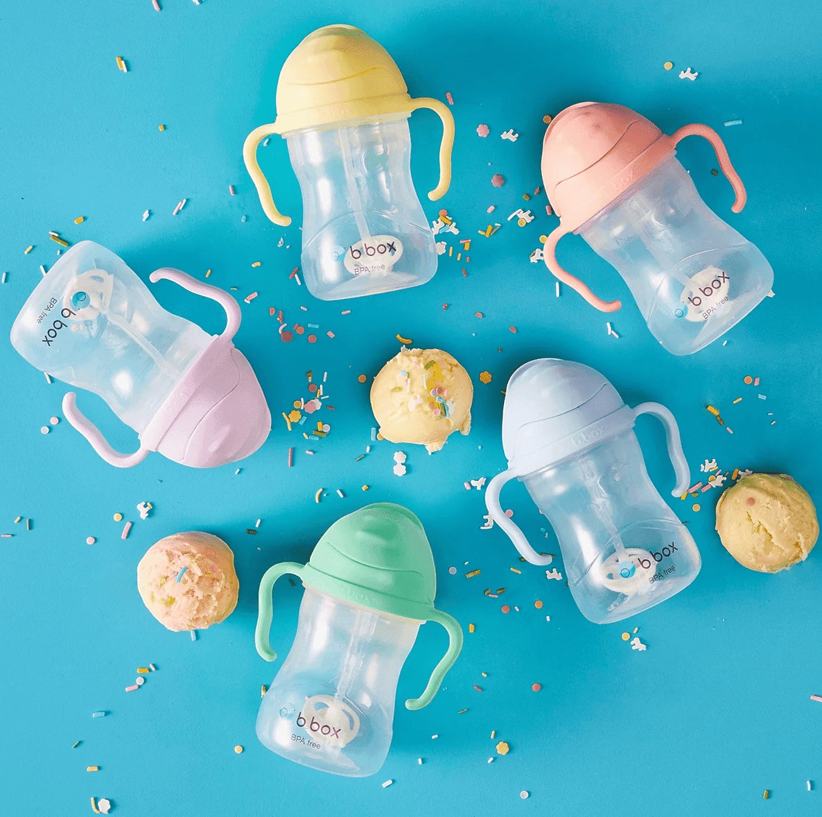 Eclectic b.box weighted straw sippy cups in various pastel colours arranged on a blue background with sprinkles.