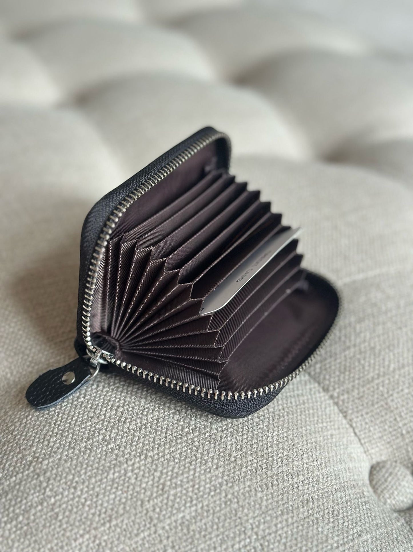 Eclectic black pebbled leather zip-around card holder with an open accordion-style interior on a tufted surface.